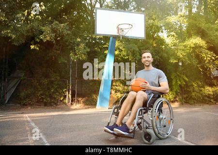 Sporty young man with ball sitting in wheelchair outdoors Stock Photo