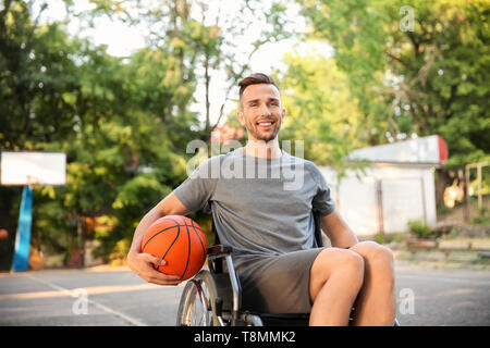 Sporty young man with ball sitting in wheelchair outdoors Stock Photo