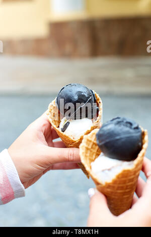 Female hand holding two charcoal and vanila ice cream in waffle cone on city background. Italian traditional ice cream gelato. Copy space. Stock Photo