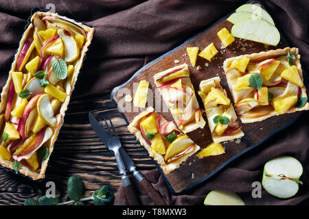 freshly baked pineapple and apple sweet tart in rectangular tart pan and the one sliced on a cutting board on an old wooden table with brown cloth, ho Stock Photo