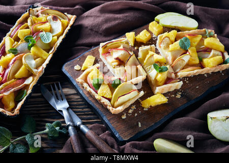 freshly baked pineapple and apple sweet tart in rectangular tart pan and the one sliced on a cutting board on an old wooden table with brown cloth, ho Stock Photo