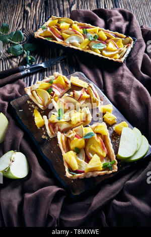 freshly baked pineapple and apple sweet tart sliced on a cutting board on an old wooden table with brown cloth, vertical view from above Stock Photo