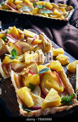 freshly baked pineapple and apple sweet tart sliced on a cutting board on an old wooden table with brown cloth, vertical view from above Stock Photo