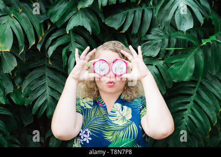 Blonde Caucasian woman holding slices of Pitaya dragon fruit in front of her eyes, kissing. Tropical leaves background, copy space. Stock Photo
