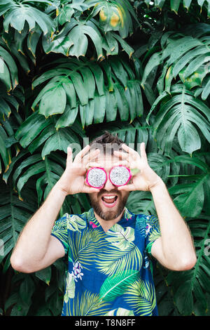 Young bearded man holding slices of Pitaya dragon fruit in front of his eyes, laughing. Tropical leaves background, copy space, vertical orientation. Stock Photo
