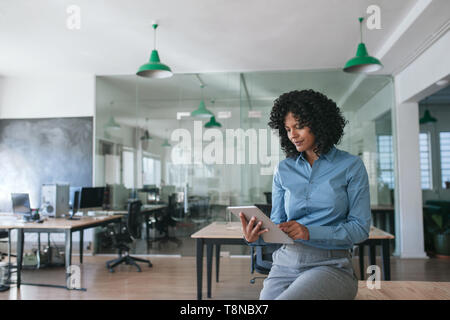 Smiling businesswoman sitting on an office table using a tablet Stock Photo