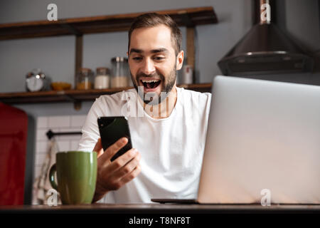 Happy young man using laptop computer while sitting at the kitchen table, holding mobile phone Stock Photo