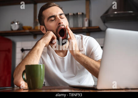 Bored young man using laptop computer while sitting at the kitchen table Stock Photo