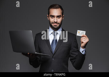 Portrait of a confident handsome businessman wearing suit isolated over black background, holding laptop computer, showing credit card Stock Photo