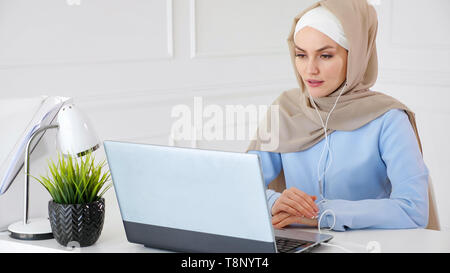Portrait of young muslim woman in hijab and traditional clothes is learning english in earphones online using computer sitting at table. Stock Photo