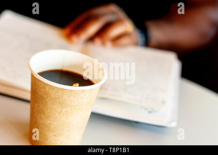 A mug of coffee with a black man reading a book in the background. Taking a break with a coffee Stock Photo