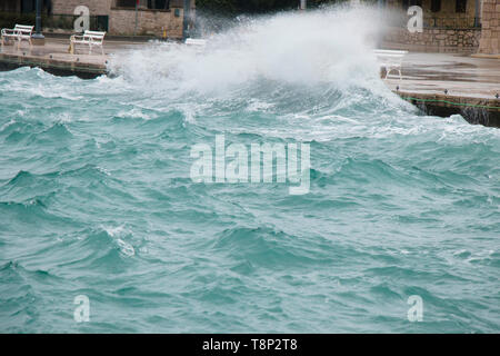 Stormy sea splashing the waves on coast promenade with benches in Dalmatia seaside town in off season during strong south wind with rain Stock Photo