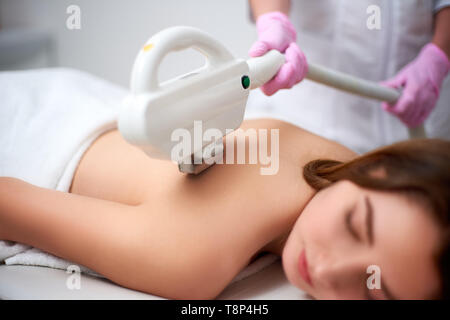 Young pretty smiling woman getting back laser epilation in beauty salon. Cosmetologist doing depilation treatment with elos device. Beautician hands i Stock Photo