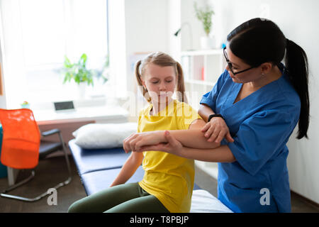 Caring long-haired nurse in blue uniform asking little girl about feelings Stock Photo