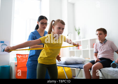 Attentive long-haired Asian therapist changing posture of little girl Stock Photo