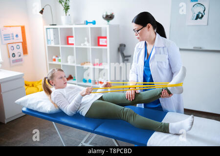 Diligent skinny little girl lying on the daybed and working with elastic rope Stock Photo