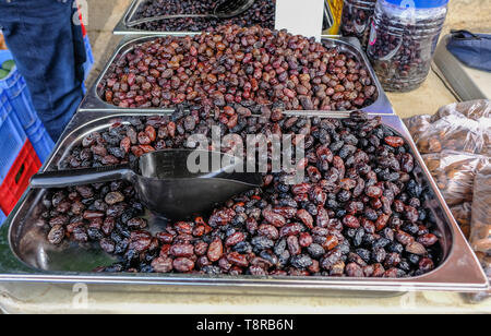 Metal containers full of black olives on a stall at a street market. Black scoop for serving these dried black olives. Stock Photo