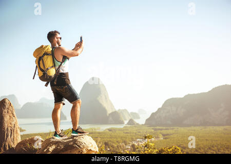 Backpacker tourist stands on big rock and takes mobile photo at viewpoint Stock Photo