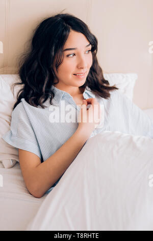 Lovely young female with calm expression, wearing blue striped shirt, sitting on white bedclothes in bedroom holding pillow, looking away, feeling rel Stock Photo