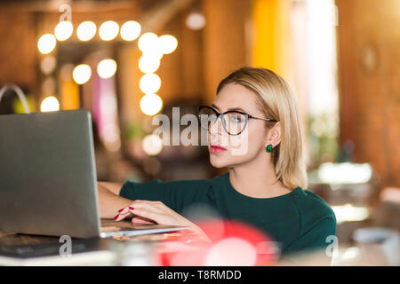 Indoor close up portrait of attractive blonde concentrated freelancer, administrator, manager in big glasses using laptop computer. Selective focus. B Stock Photo