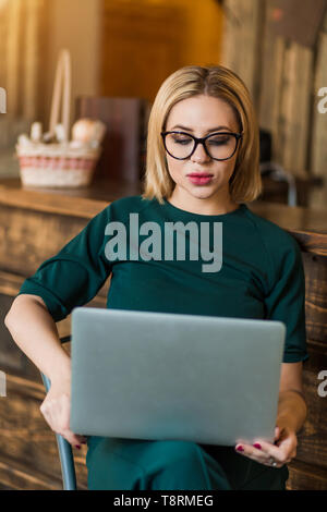 Confident female freelancer sitting on chair holding laptop looking with serious and thoughtful expression. Young businesswoman in glasses using PC fo Stock Photo