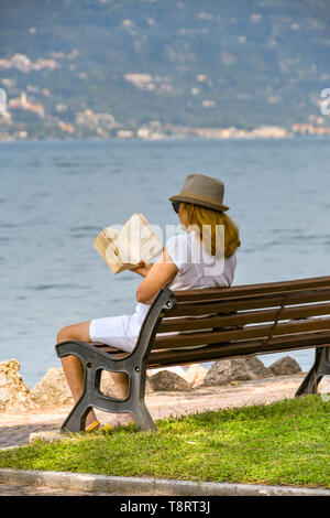 TORRI DEL BENACO, LAKE GARDA, ITALY - SEPTEMBER 2018: Person reading a book sitting on a bench lakeside in the town of Torri del Benaco on Lake Garda Stock Photo