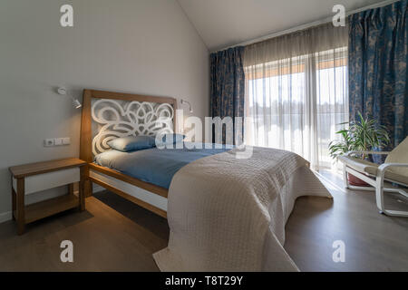Main bedroom with big original bed, bedside tables, lamps on the wall, white armchair and flower pot near the window with curtains in the modern house Stock Photo