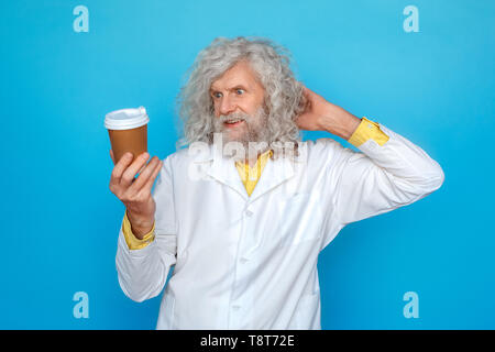 Old long-haired man wearing doctor's gown studio isolated on blue wall standing looking at cup of hot coffee scratching head smiling confused Stock Photo