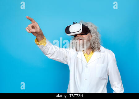 Old long-haired man wearing doctor's gown and virtual reality headset studio isolated on blue wall standing pointing forward smiling excited Stock Photo