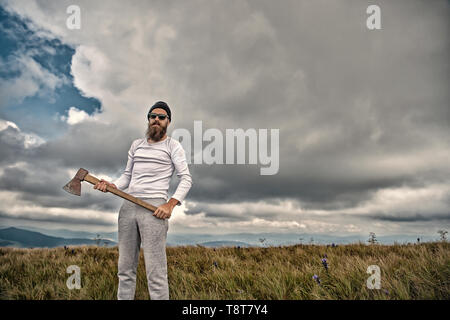 Hipster bearded man in sunglasses with axe stand on mountain landscape on cloudy sky. Logging and chopping concept. Wanderlust, vacation, travelling, vintage filter Stock Photo