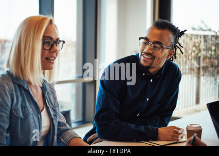 Two diverse young business colleagues smiling while having a meeting around a desk in a modern office Stock Photo