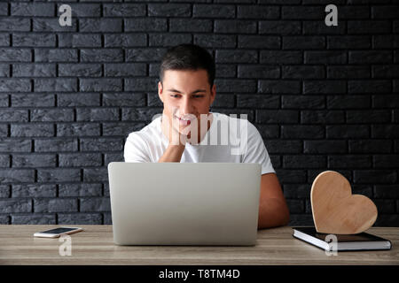 Young man having online dating while sitting at table with laptop against brick wall Stock Photo