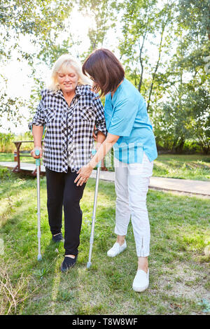 Nursing woman helps senior woman with crutches while walking learning in rehab Stock Photo