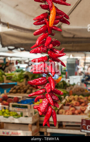 Red hot spicy chili peppers hanging on a string at a farmers market in Rome. Apples and other fresh fruits and vegetables are for sale in the backgrou Stock Photo