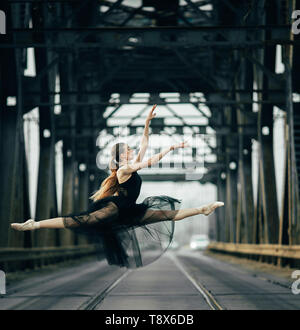 Ballerina jumping in twine pose in a black transparent dress on the road and rails against the background of metal supports. Stock Photo