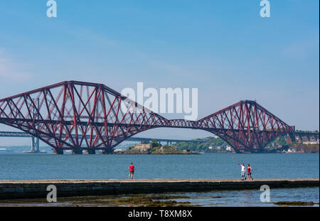 South Queensferry, Scotland, United Kingdom, 15 May 2019. UK Weather: People walk along the pier on a sunny day on the Firth of Forth coast at the iconic Forth Rail Bridge and Forth road bridge in distance Stock Photo