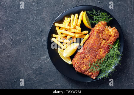Breaded hake fillet served on a black plate with chips, fresh dill and lemon slices, view from above, flatlay, copy space Stock Photo