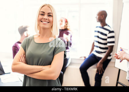 Smiling young female designer standing confidently with her arms crossed after a boardroom meeting with colleagues standing in the background