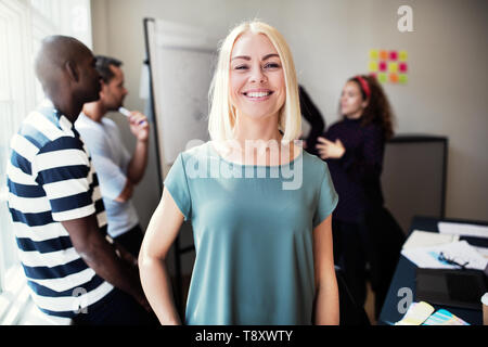 Young female designer smiling while standing in an office after a meeting with colleagues working together in the background Stock Photo