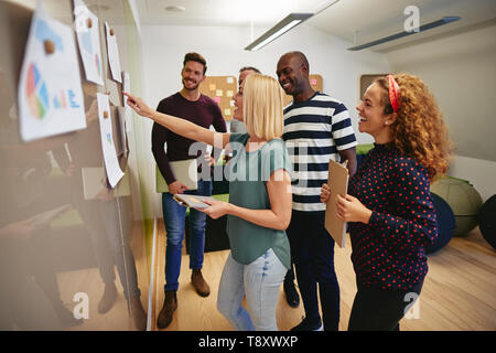 Diverse group of smiling colleagues looking at graphs and charts taped to a wall while having a meeting together in a modern office Stock Photo