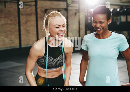 Two young women in sportswear laughing while standing together in a gym with a skipping rope after working out Stock Photo