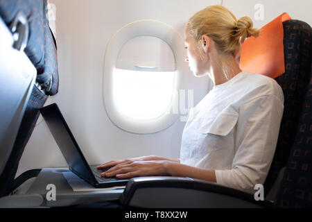 Attractive caucasian female passenger looking through the plain window while working on modern laptop computer using wireless connection on board of Stock Photo