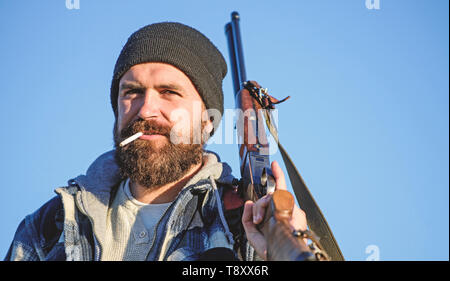 Hunting masculine hobby concept. Man brutal bearded guy gamekeeper blue sky background. Brutality and masculinity. Hunter with rifle gun close up. Guy bearded hunter spend leisure hunting and smoking. Stock Photo