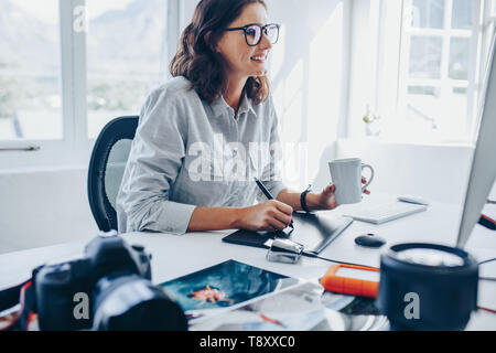 Young woman sitting at desk editing images on computer. Female photographer retouching photos in office using graphic tablet and digital pen. Stock Photo