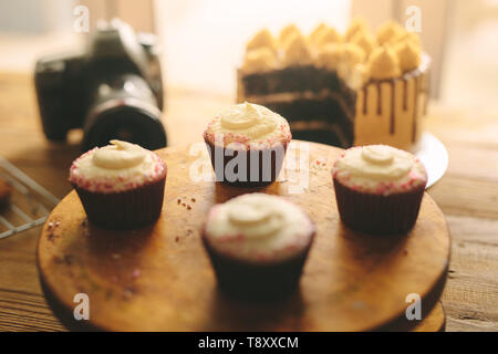 Cupcakes on wooden board with cake and camera on table at the back. Stock Photo