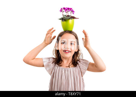 Girl carrying flower pot on head, having fun smiling. Little girl in dress holding green flower pot with viola flowers on her head having fun, goofing Stock Photo