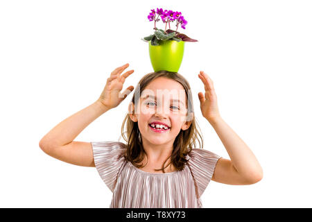 Girl carrying flower pot on head, having fun smiling. Little girl in dress holding green flower pot with viola flowers on her head having fun, goofing Stock Photo