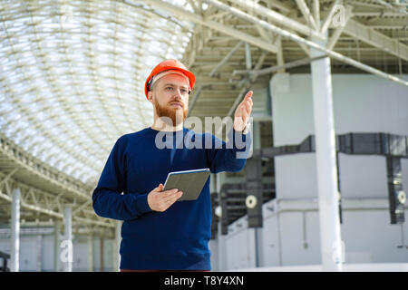 The average plan portrait of an adult man in a helmet talking, hand pointing to the side, holding a tablet PC, horizontal, space for signature Stock Photo