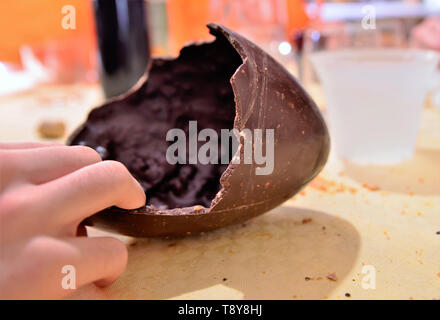 An Easter egg has been opened, the broken chocolate shell is on the table resting in its wrapping. A Caucasian man's hand cuts off a piece of food. 30 Stock Photo