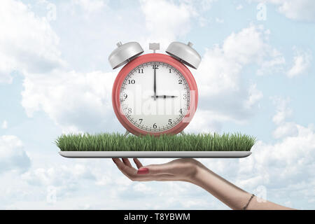 Female hand holding red alarm clock on green grass model on blue sky background Stock Photo